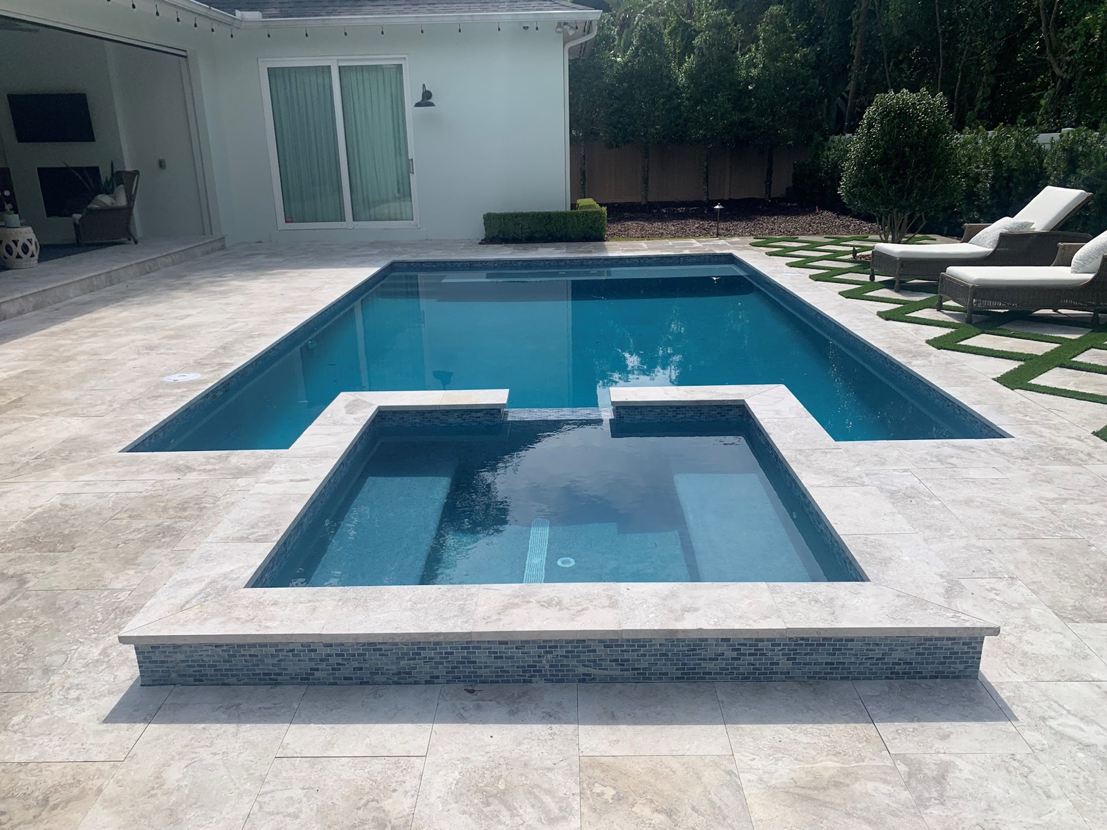 Rectilinear pool with attached spa, travertine paver deck, and loungers on turf inlay strip, white single-story home