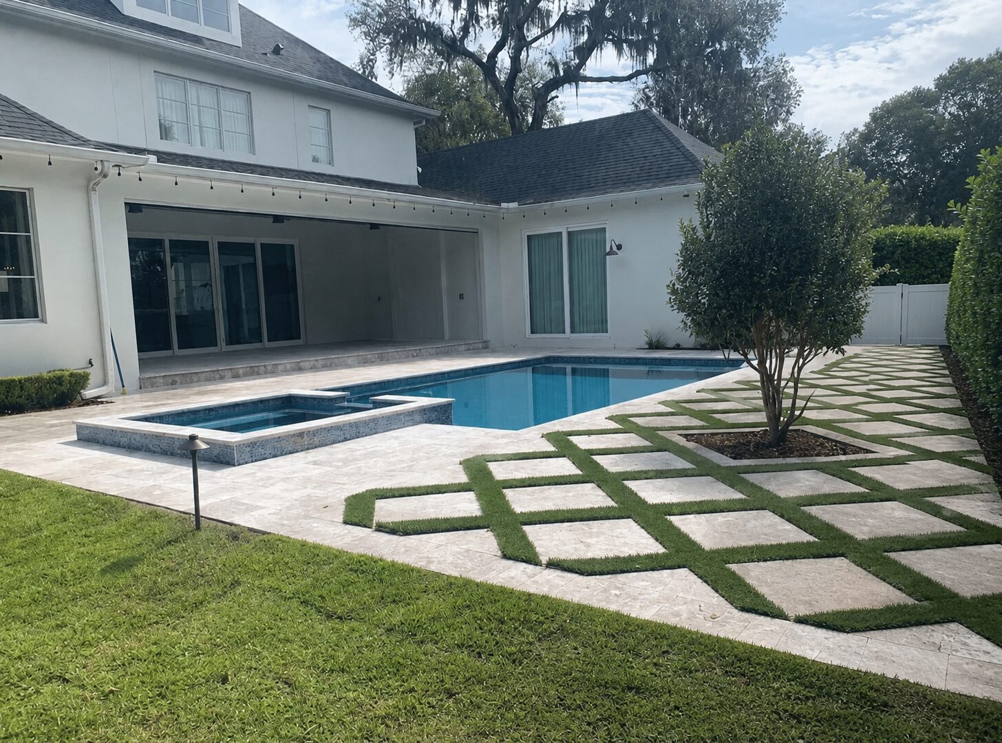 Pool and raised spa with travertine decking and geometric turf inlays, two-story white home in background